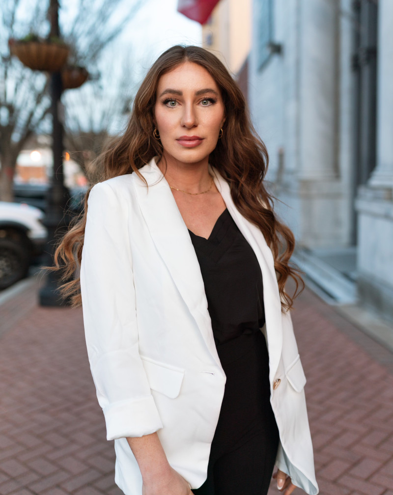 Kate Nemec standing on a brick sidewalk in a white blazer during an outdoor professional portrait.